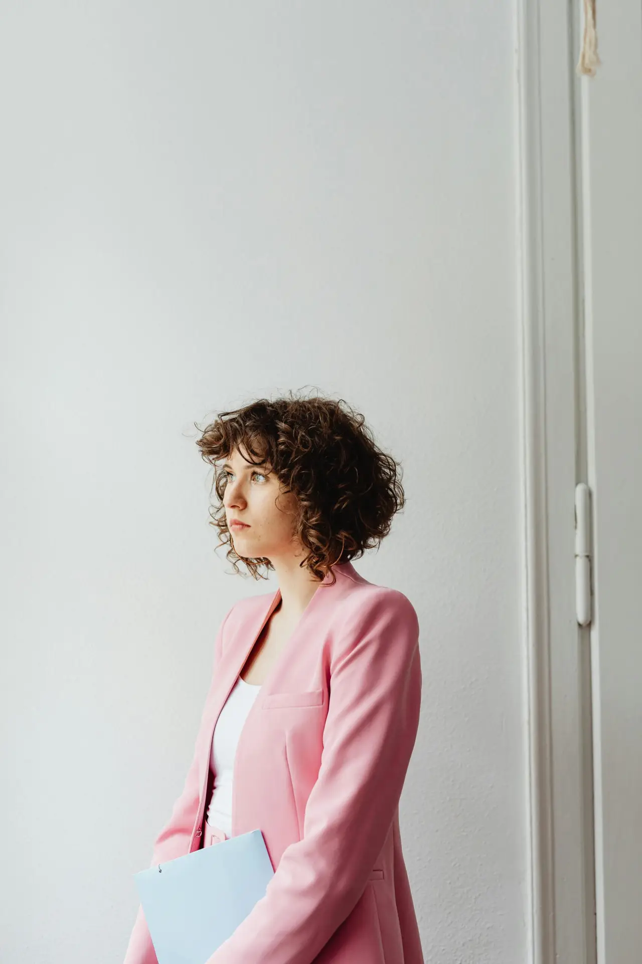 Young woman in a pink blazer holding a folder, standing indoors with an introspective look.